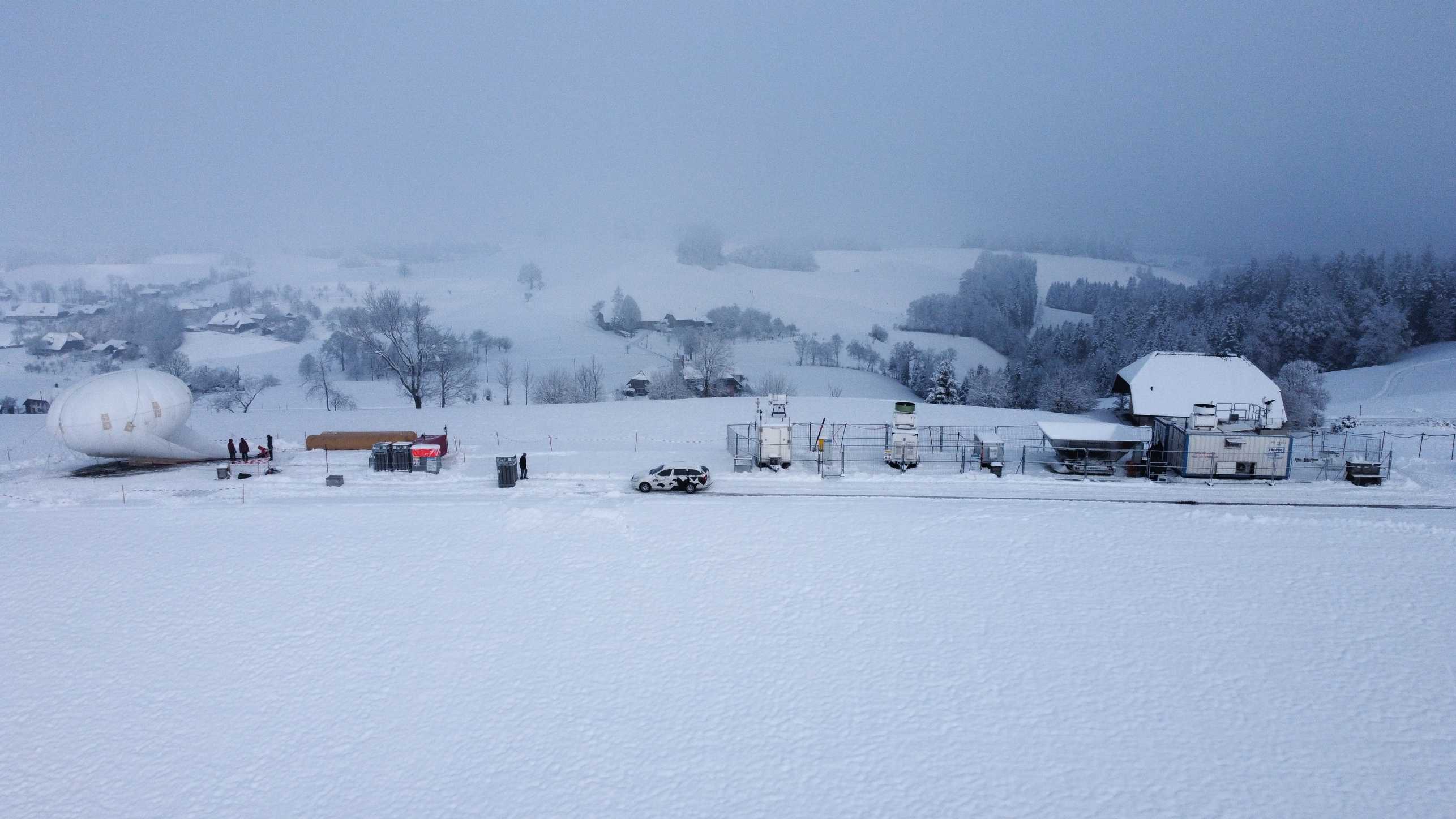 Vergrösserte Ansicht: Alle Geräte die in CLOUDLAB betrieben werden nebeneinander. Von links nach rechts: HoloBalloon, Trailer mit Mikrowellenradar, Parsivel und bodenbasierte Aerosolmessungen, Ka band Wolkenradar, Windradar mit Trailer (Winter 2022/2023), zwei TROPOS Container mit ebenfalls Parsivel, Mikrowellenradar und Ka Band Wolkenradar als auch Windlidar, Dual Wellenlängen Lidar und zuletzt W Band Wolkenradar.